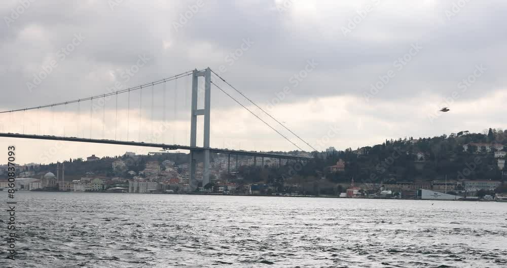 Large modern bridge with moving automobiles across wide river in city. Seagull flies over surface of rippling water on cloudy summer day before rain slow motion