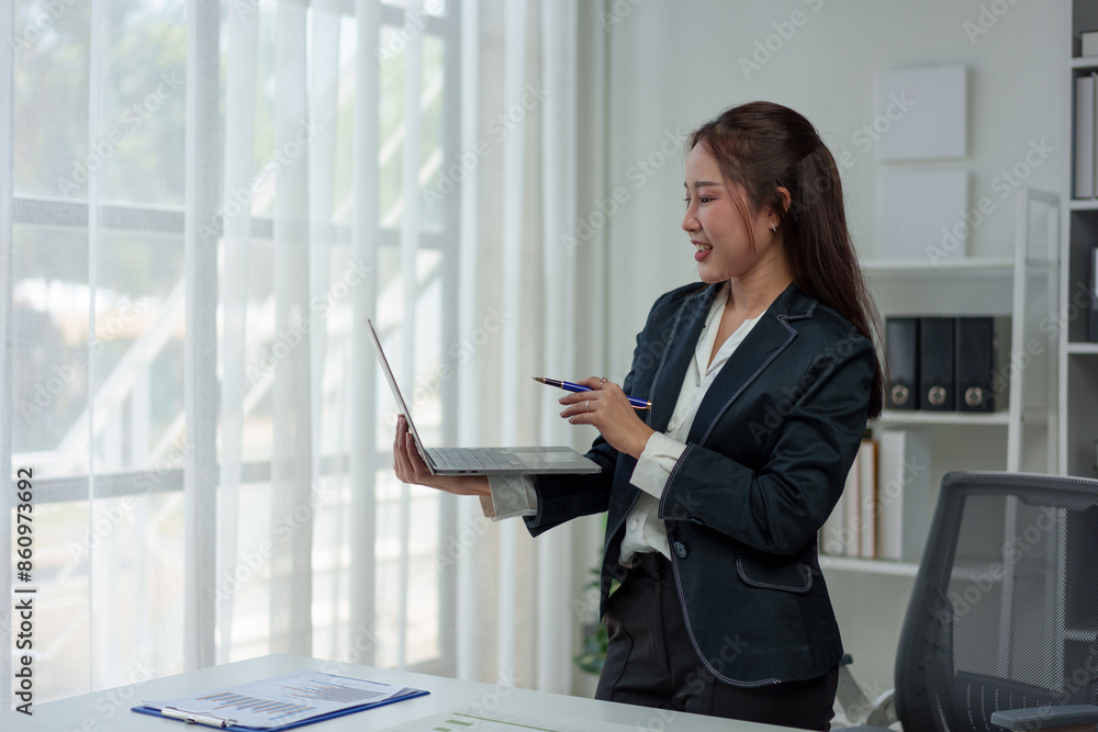 Cheerful and confident Asian businesswoman stands presenting bar chart data from a laptop folder in an online meeting in the office. Startup business ideas, accounting, online marketing. Cheerful and confident Asian businesswoman stands presenting bar chart data from a laptop folder in an online meeting in the office. Startup business ideas, accounting, online marketing.