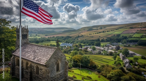 An American flag flying high above a historic stone church, with a charming village and rolling hills stretching out below