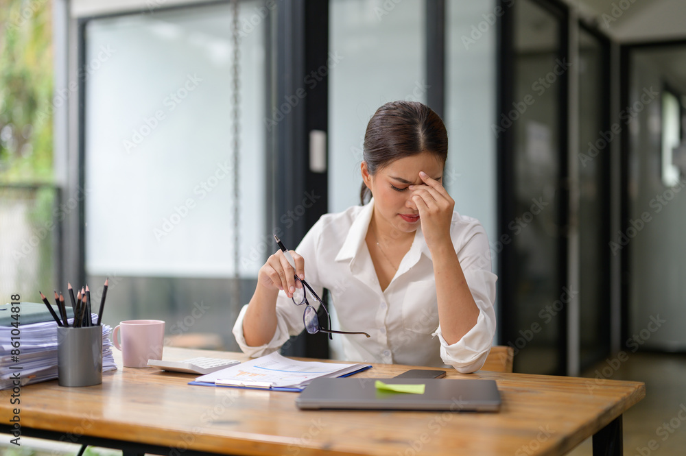 Tired Asian woman working with tax documents on laptop computer in office. Sad, worried, depressed or stressed employee life. Stretch your muscles due to sitting and working for a long time. Tired Asian woman working with tax documents on laptop computer in office. Sad, worried, depressed or stressed employee life. Stretch your muscles due to sitting and working for a long time.