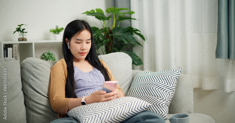 Portrait of Relaxed young Asian woman wear wireless headphones listen to music while sitting playing mobile phone on the sofa in the living room at morning ,Free time Portrait of Relaxed young Asian woman wear wireless headphones listen to music while sitting playing mobile phone on the sofa in the living room at morning ,Free time