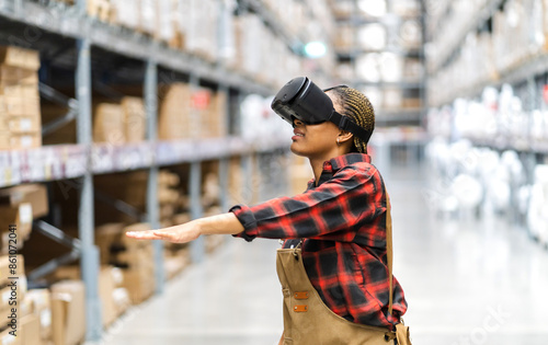 Young africn woman wearing virtual reality (VR) goggles stands in middle of warehouse with shelves stocked, traditional workwear and modern technology, technology logistics in industrial and warehouse photo