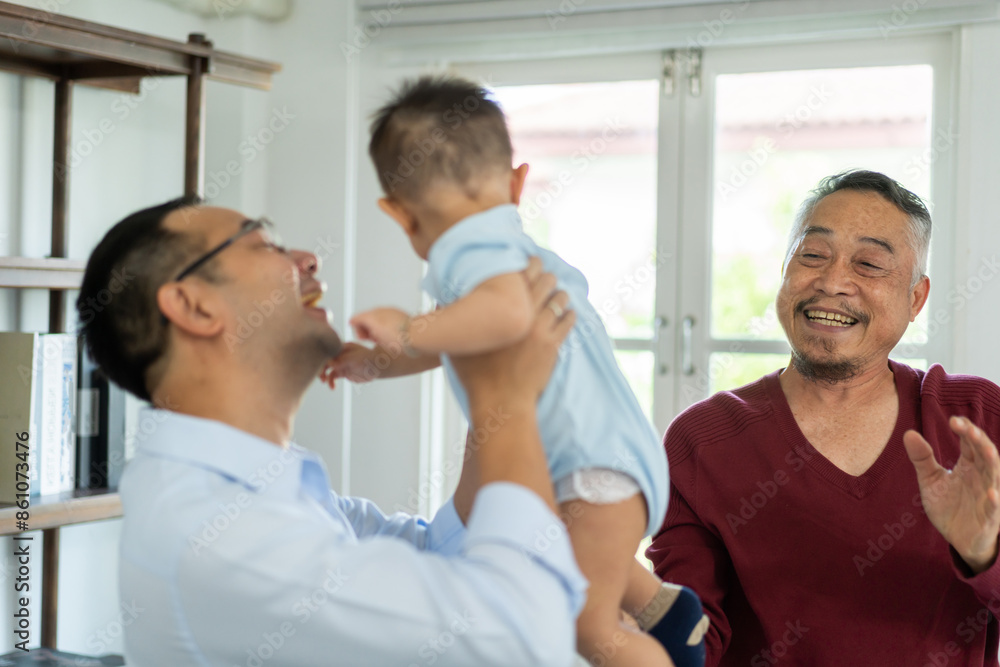 Obraz premium Joyful moment of multigenerational family at home, father holding up his baby smiling and playing while the grandfather looks on with a big smile enjoying the happy interaction, love within a family