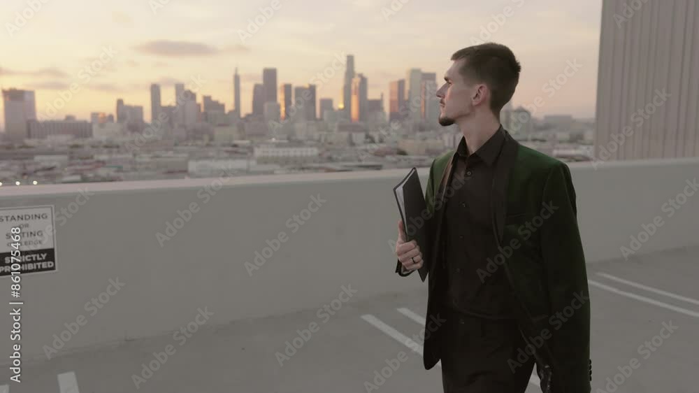 A successful young businessman, dressed in a stylish dark green suit, is standing on a rooftop parking structure with a laptop. He gazes contemplatively at the city skyline at sunset A successful young businessman, dressed in a stylish dark green suit, is standing on a rooftop parking structure with a laptop. He gazes contemplatively at the city skyline at sunset