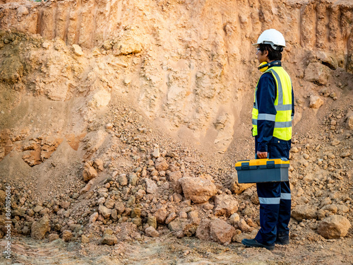 Geotechnical engineering concept. Male soil engineer in protective suit with reflective green vest and helmet holding toolbox while inspecting laterite soil for construction improvement base road work