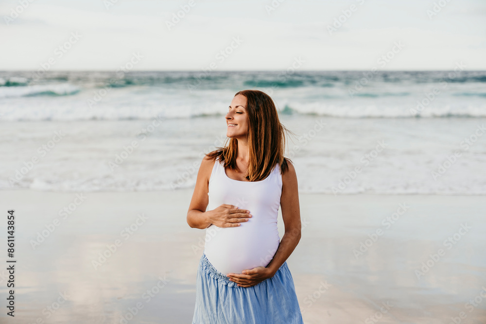 Smiling pregnant woman with hands on stomach standing near sea