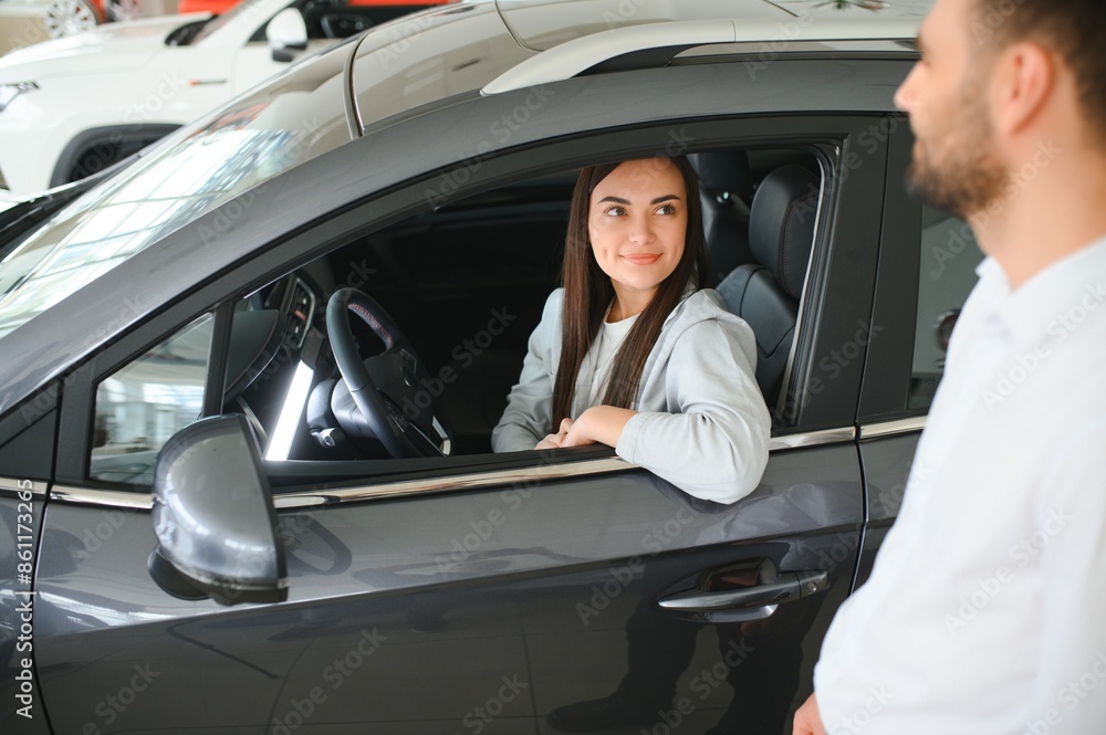 Satisfied happy caucasian female client customer woman sitting at the wheel of new car while male shop assistant helping her choose it Satisfied happy caucasian female client customer woman sitting at the wheel of new car while male shop assistant helping her choose it