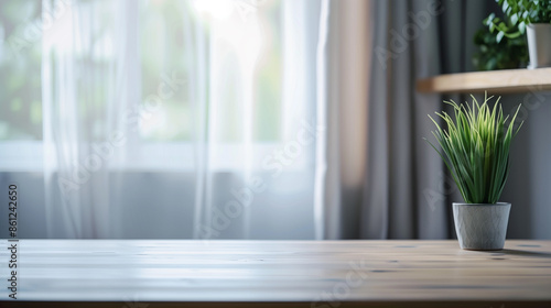 The blurred empty table top in the living room had a light grey curtain and window behind it. A modern interior design was shown with a blurred green plant on the side of the wall and a wooden shelf 