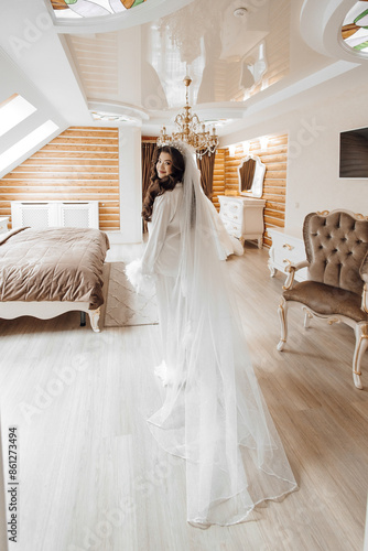 A woman in a white dress stands in front of a bed with a white veil on her head