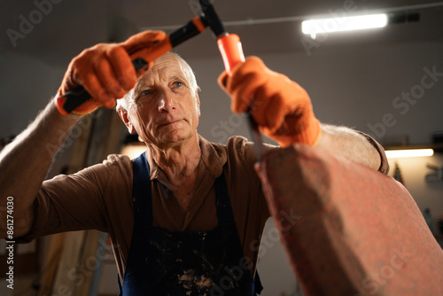 An elderly man in gloves and a blue apron begins to restore an old chair in a workshop. holds a hammer removes old fabric. concept second life of old furniture. Copy space. photo