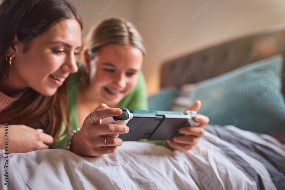 Two Teenage Girls At Home In Bedroom Together Playing On Handheld Computer Gaming Device Together Two Teenage Girls At Home In Bedroom Together Playing On Handheld Computer Gaming Device Together