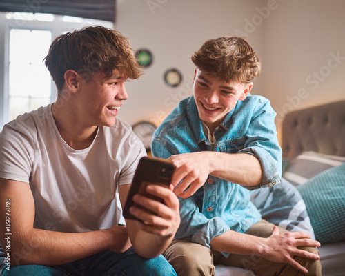 Two Teenage Boys At Home In Bedroom Looking At Social Media On Mobile Phone And Laughing