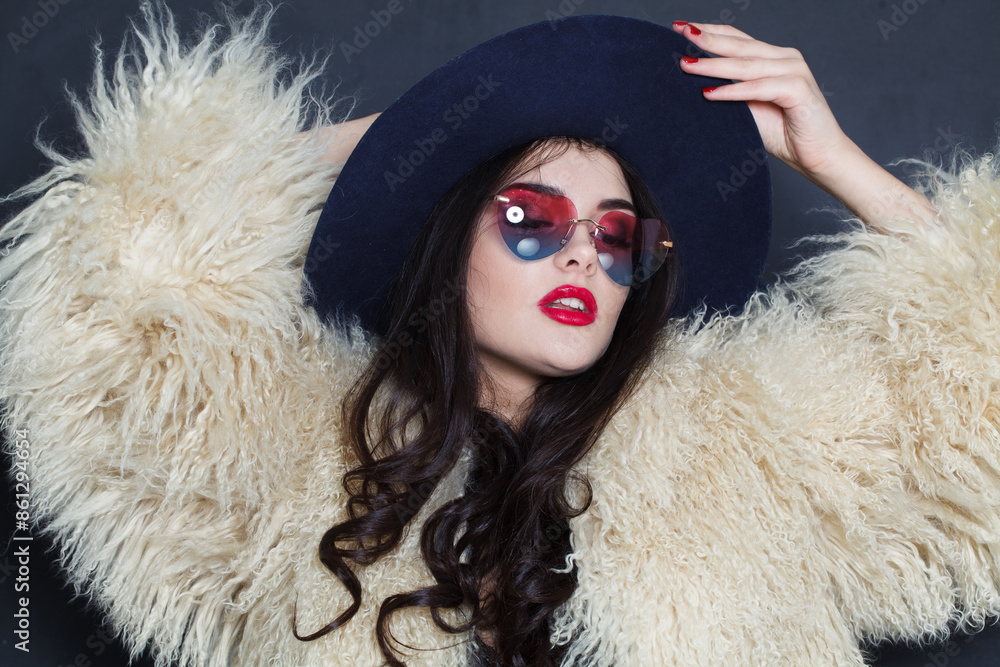 Charming young adult woman fashion model with bright make-up and long wavy hair wearing blue hat and sunglasses posing against blue studio wall background Charming young adult woman fashion model with bright make-up and long wavy hair wearing blue hat and sunglasses posing against blue studio wall background