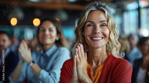 A smiling woman in an orange blouse claps her hands in a modern office, surrounded by a diverse group of colleagues, creating a warm and inclusive atmosphere.