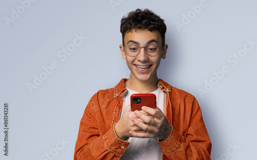Smiling funny curly hair young man wear braces, glasses, orange denim shirt hold typing text look read cell phone cellular smartphone cellphone, isolated grey gray background.