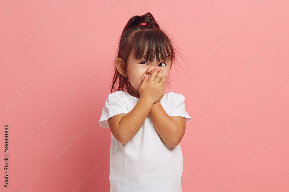 Little Asian girl covers her mouth with her hand with her eyes wide open in shame, said something superfluous and inappropriate, expresses embarrassment and shyness while standing on a pink isolated. Little Asian girl covers her mouth with her hand with her eyes wide open in shame, said something superfluous and inappropriate, expresses embarrassment and shyness while standing on a pink isolated.
