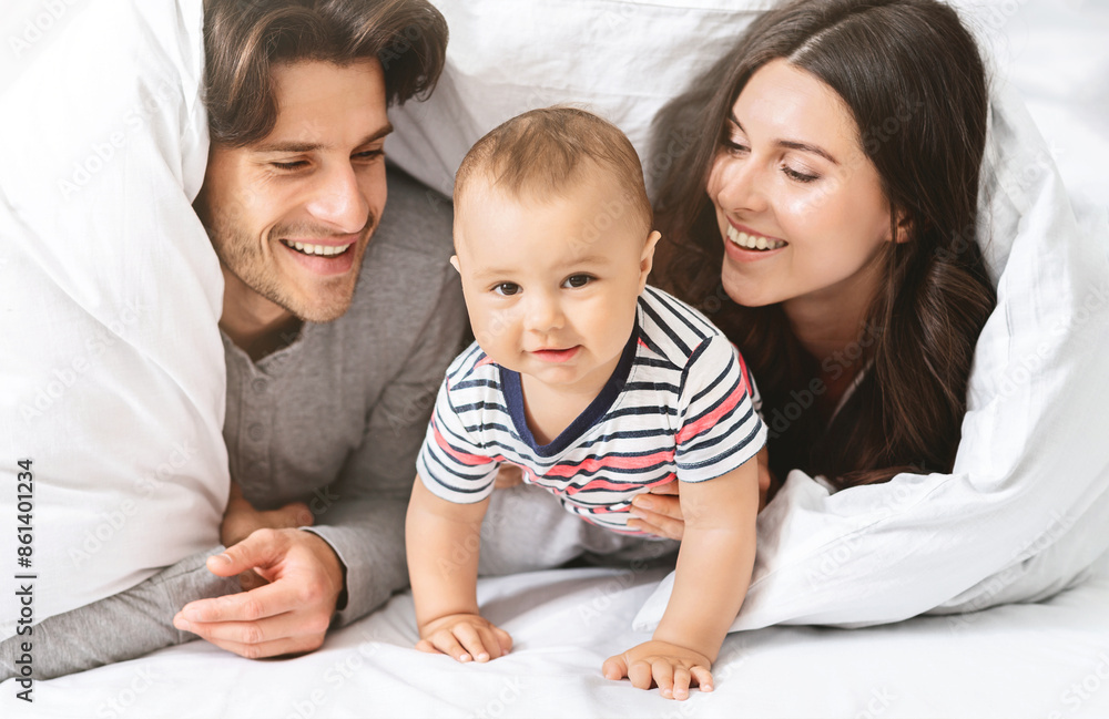 A happy family of three is playing under the covers in bed. The baby is crawling and looking at the camera while the parents are smiling and looking at their child. A happy family of three is playing under the covers in bed. The baby is crawling and looking at the camera while the parents are smiling and looking at their child.