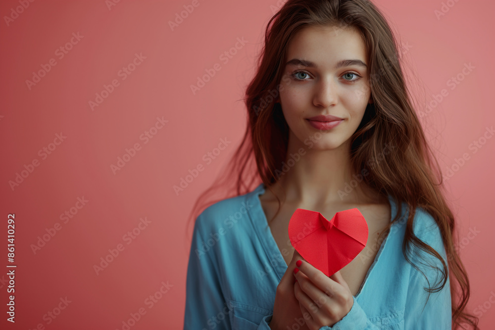 Young woman holding a red paper heart, symbolizing love and emotion, standing against a pink background with a serene expression Young woman holding a red paper heart, symbolizing love and emotion, standing against a pink background with a serene expression