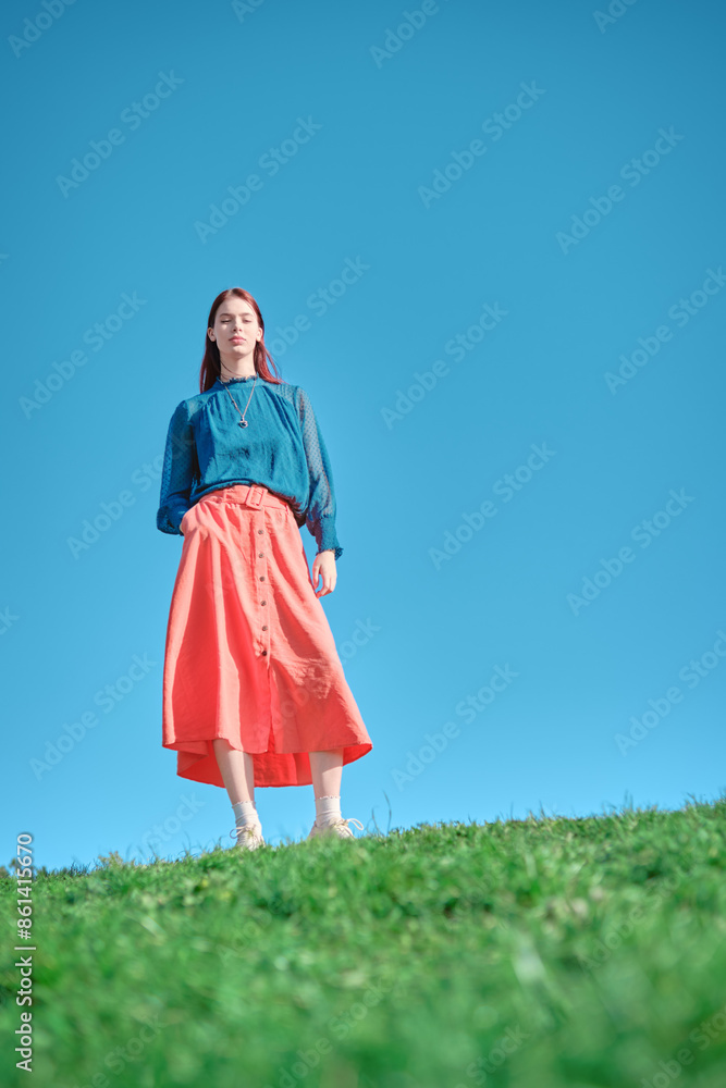 Teen girl in blue blouse and coral skirt standing on green grass with one hand in pocket against clear blue sky, low angle shot Teen girl in blue blouse and coral skirt standing on green grass with one hand in pocket against clear blue sky, low angle shot