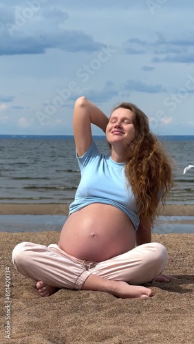 Happy young pregnant woman on the beach sitting on the sand and stroking her belly