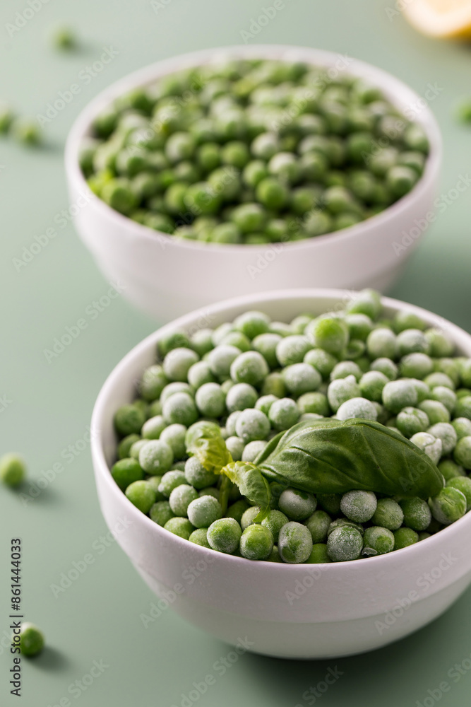 Two white bowls filled with frozen green peas, with a fresh basil leaf on a green background. Two white bowls filled with frozen green peas, with a fresh basil leaf on a green background.