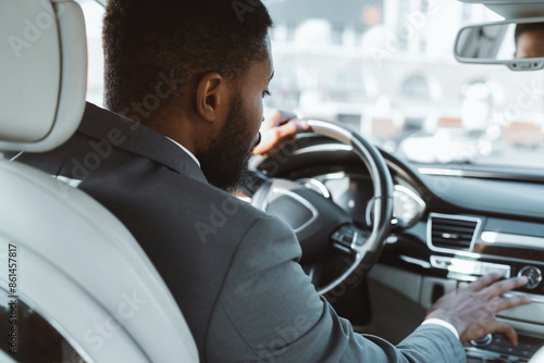 A man in a suit is driving a car, his hand resting on the dashboard. The man is focused on the road ahead, his expression serious. The city can be seen in the background © Prostock-studio