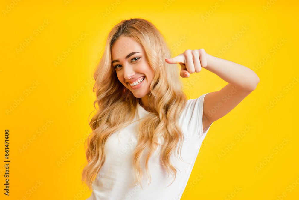 A young woman with long blonde hair is smiling and pointing at the camera. She is wearing a white t-shirt and is standing in front of a yellow background. The image conveys a sense of confidence A young woman with long blonde hair is smiling and pointing at the camera. She is wearing a white t-shirt and is standing in front of a yellow background. The image conveys a sense of confidence