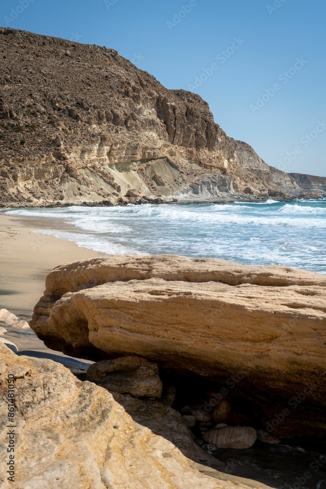 Fototapeta premium Scenic view of a rocky beach with waves crashing on the shore under a clear blue sky.