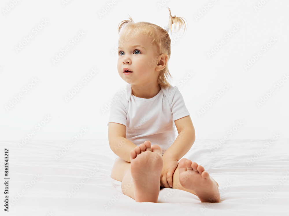 Curious kid, baby girl with blonde pigtails sits on white surface, dressed in onesie against white studio background. Concept of childhood, motherhood, care, life, birth. Copy space. Curious kid, baby girl with blonde pigtails sits on white surface, dressed in onesie against white studio background. Concept of childhood, motherhood, care, life, birth. Copy space.