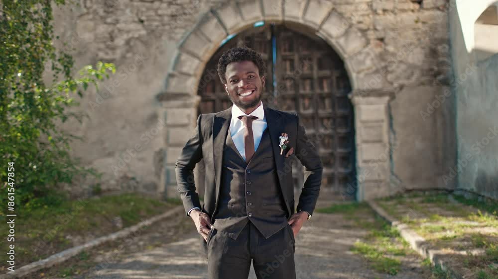 Happy African American groom standing in front of entrance to old castle or church and positively smiling at camera. Handsome man with elegant black suit waiting to start wedding ceremony outside. Happy African American groom standing in front of entrance to old castle or church and positively smiling at camera. Handsome man with elegant black suit waiting to start wedding ceremony outside.