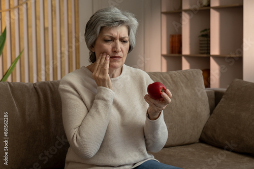 A woman sits on a couch in her living room and take a bite of an apple. She grimaces in pain, likely experiencing a toothache. Depicts the discomfort associated with tooth pain