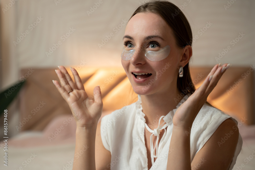 A young woman applies eye patches in her bedroom at night. She is sitting on the edge of a bed with a plant in the background A young woman applies eye patches in her bedroom at night. She is sitting on the edge of a bed with a plant in the background