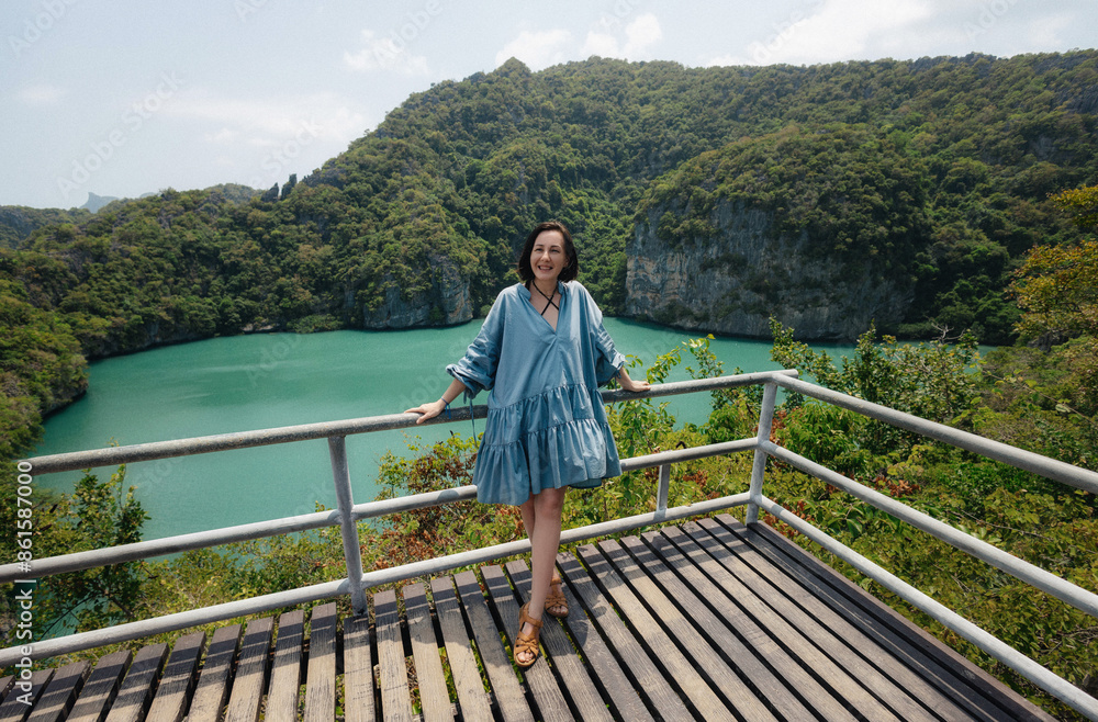 Fototapeta premium A woman wearing a blue dress leans on a railing and enjoys a view of a scenic lagoon surrounded by lush green hills on a sunny day