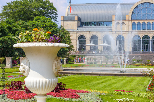 White vase with flowers in foreground, green grass, fountain with water jets, greenhouse building of Flora Botanical Garden in blurred background, sunny spring day in Cologne, Germany