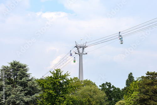 Cabins hanging from cable car cables over Rheinpark public park, metal tower and structures, green treetops, cloud-covered sky in background, cloudy day in Cologne, Germany