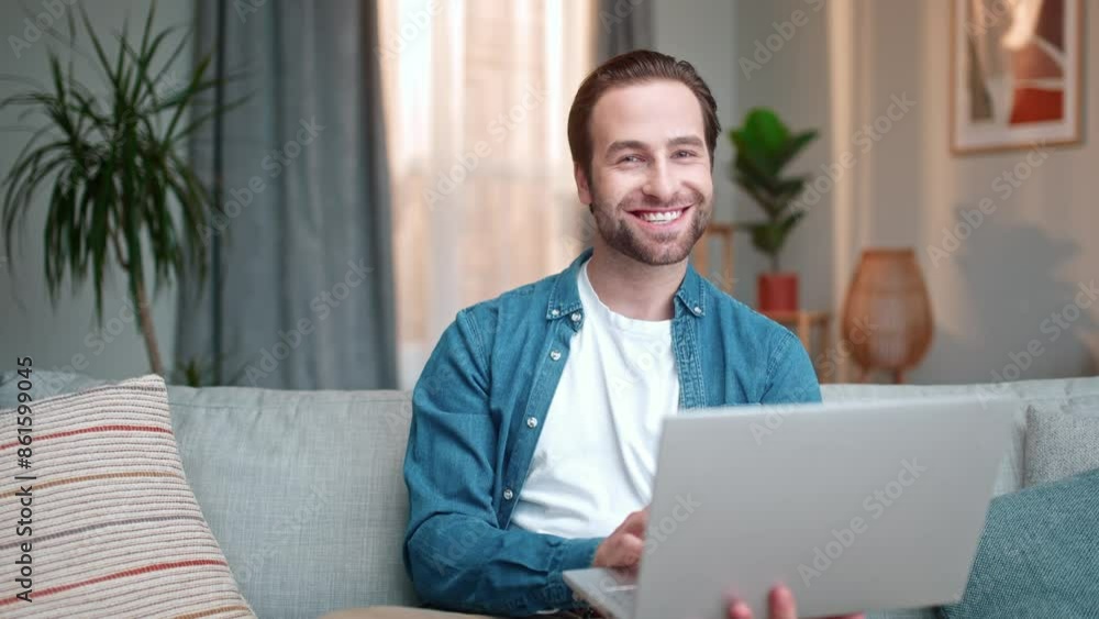 Cheerful man working with laptop sitting on sofa. Male sitting on couch and working distantly. Happy man working online. Male freelancing. Man holding laptop and smiling at camera. Cheerful man working with laptop sitting on sofa. Male sitting on couch and working distantly. Happy man working online. Male freelancing. Man holding laptop and smiling at camera.