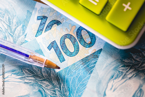 Vista superior em closeup de notas de 100 reais com uma calculadora e uma caneta na composição. Real BRL - moeda do Brasil. Impostos, imposto de renda, fundo de garantia -  fgts e salário mínimo.