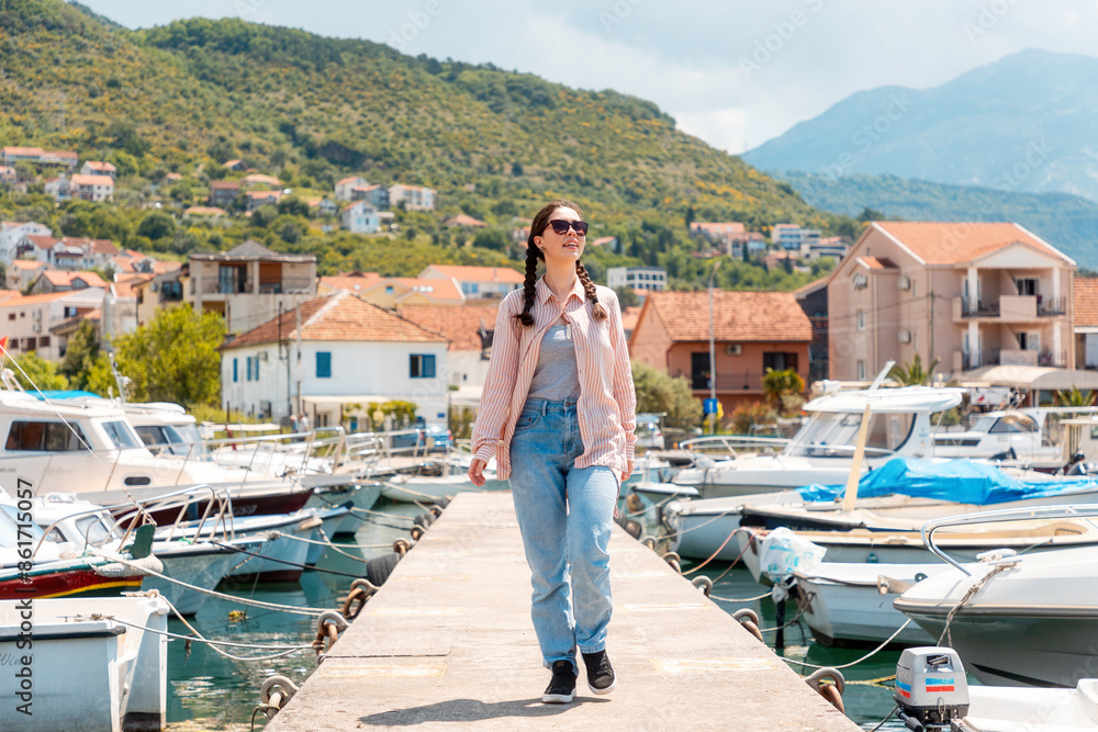 Obraz premium Happy independent Caucasian young woman walking on pier. Mediterranean village, mountains and boats in background. Summer vacation and travel