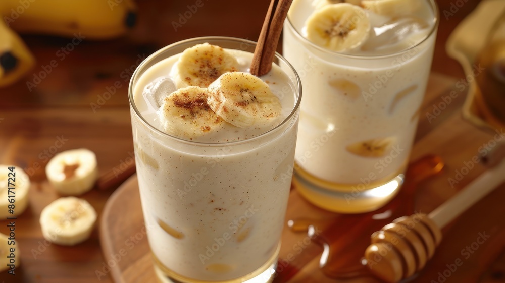 Two glasses of refreshing banana smoothie topped with banana slices and cinnamon sticks, served on a wooden table with honey. Two glasses of refreshing banana smoothie topped with banana slices and cinnamon sticks, served on a wooden table with honey.