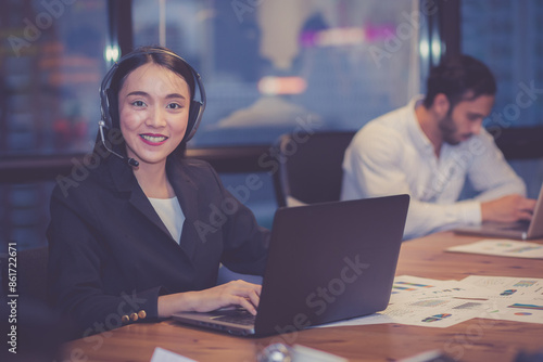 Professional young asian businesswoman wearing headset working on laptop in modern office with colleague in background, business woman wearing headphone for customer service, business concept.