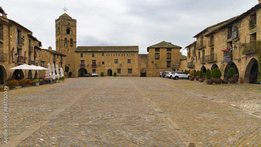 Obraz premium Una panorámica de la Plaza Mayor de Ainsa, Aragón, España, revela una plaza empedrada rodeada de edificios históricos de piedra con arcos en la planta baja. 