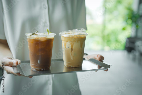 Close up and selective focus hand of employee girl holding tray of ice lychee emericano and cappuccino at cafe.