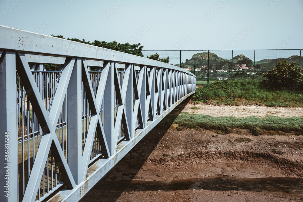 A long metal bridge extends over a riverbed, with distant hills and a fence in the background. A long metal bridge extends over a riverbed, with distant hills and a fence in the background.