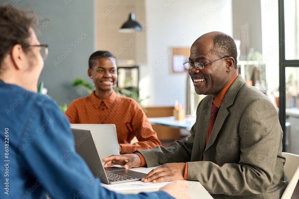 Side view portrait of Black senior man laughing enjoying work with young colleagues in office during business meeting Side view portrait of Black senior man laughing enjoying work with young colleagues in office during business meeting