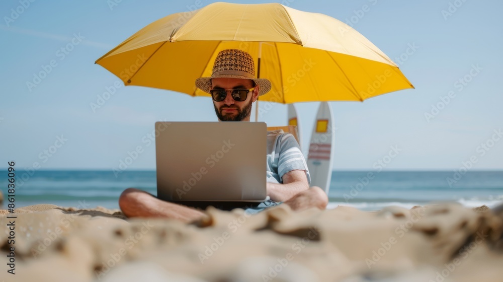 Freelancer working on a laptop under a beach umbrella with ocean view. Relaxed work environment on the beach, laptop in hand, surfboard nearby.