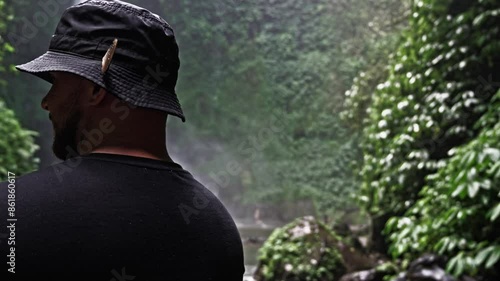 A man wearing a black outfit and bucket hat with a butterfly perched on it enjoys the view of Nungnung Waterfall, surrounded by lush green foliage. The serene natural setting highlights the beauty
