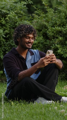 Portrait of happy young Indian man sitting in the summer park on the grass and  writes a message on a mobile phone.