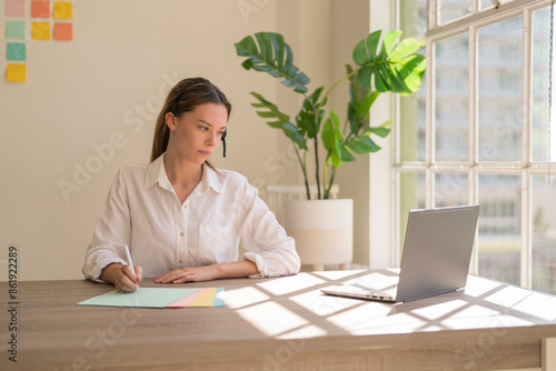 Gen Z female call center worker at desk wearing headset  and writing notes with pen, looking at computer with neutral expression