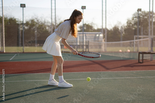 female tennis player practices hitting a bounce ball shot on a sunny day. The player is wearing a white skirt and a white shirt
