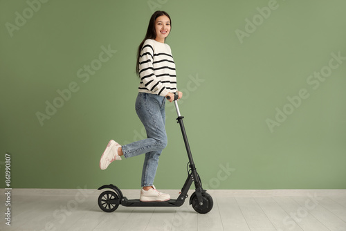 Beautiful young Asian woman riding modern electric kick scooter near green wall
